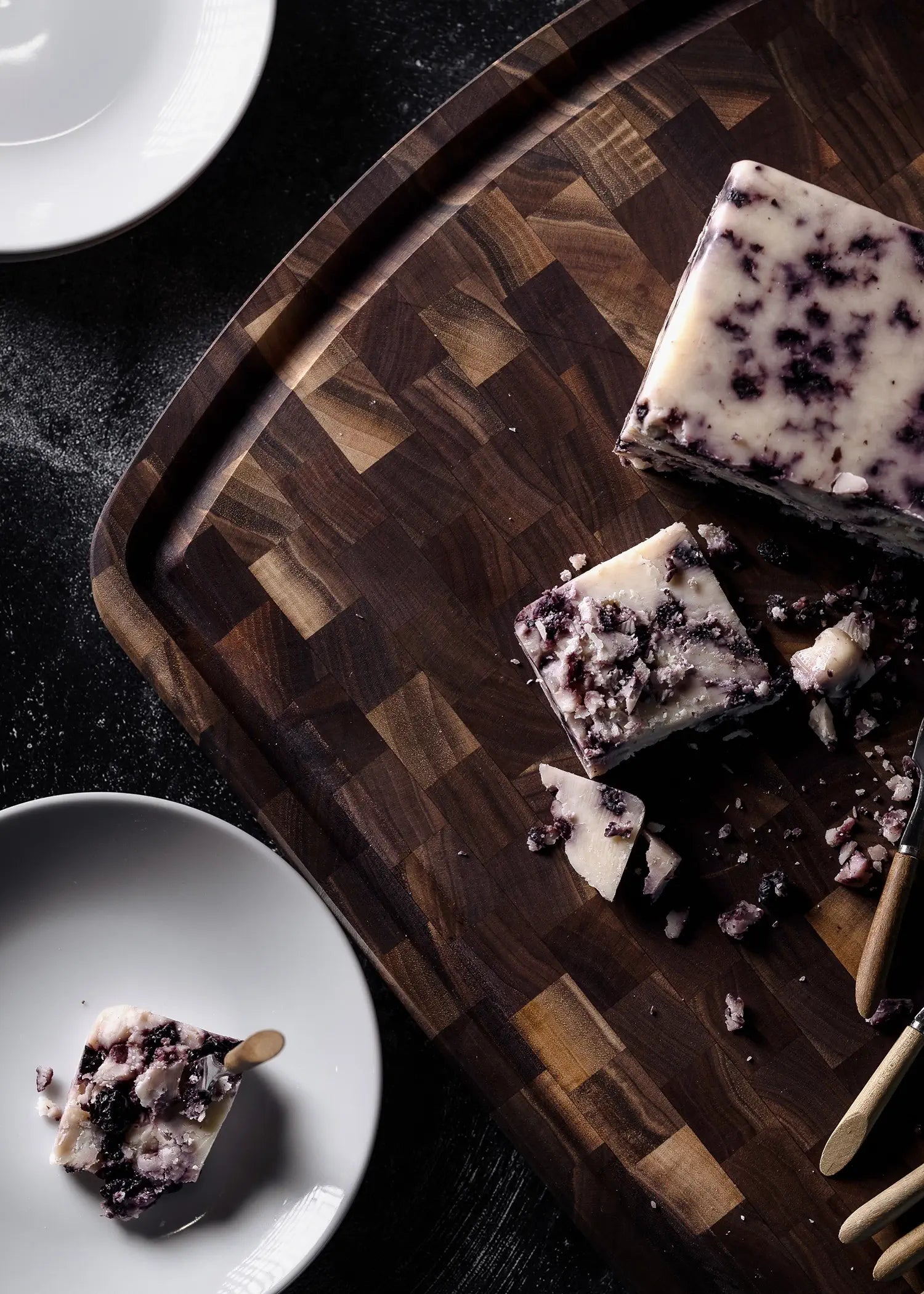 A block of crumbly cheese with dark berry-like bits sits partially sliced on a rich wooden cutting board beside a small plate.