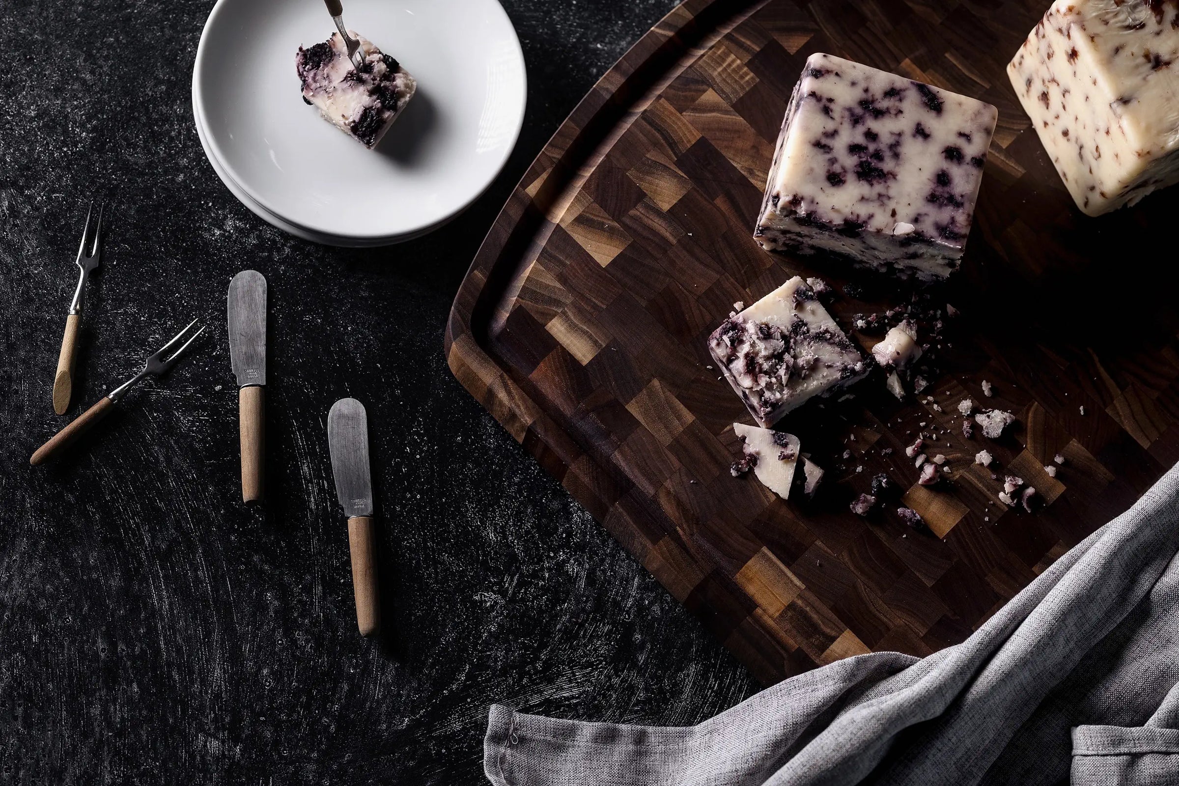 Cubes of crumbly cheese with dark berry-like specks rest on a wooden cutting board, surrounded by cheese knives, forks, and a cloth napkin on a dark textured surface.