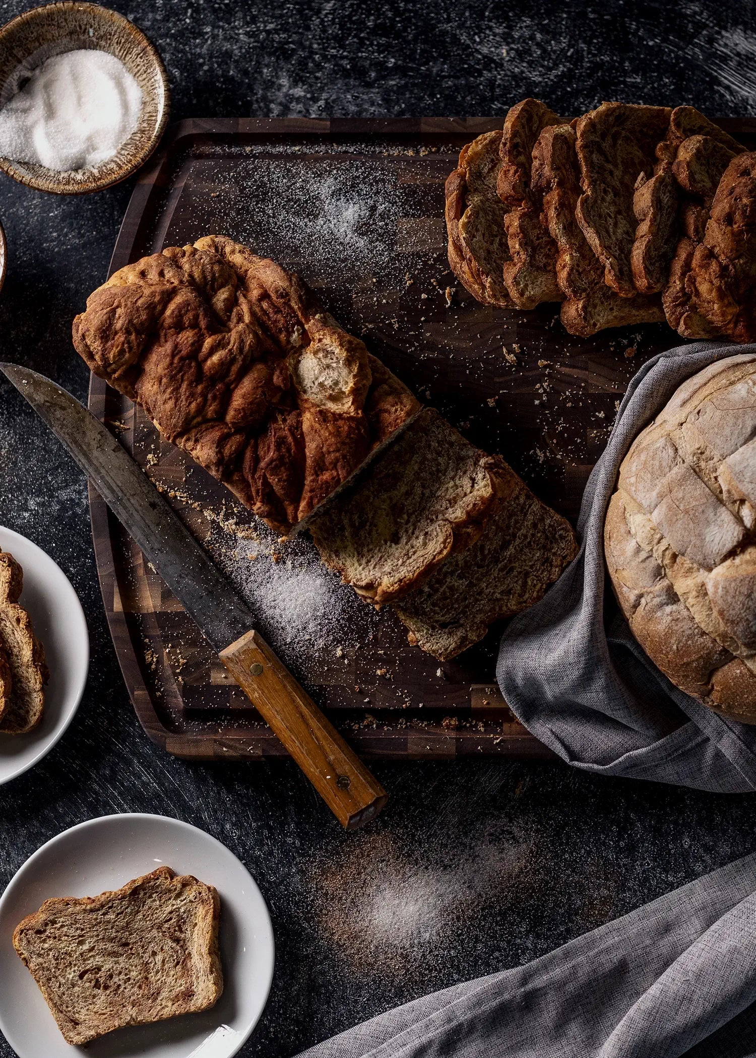 Rustic sliced bread on a dark wooden board with a knife, surrounded by plates, a round loaf, and scattered flour.