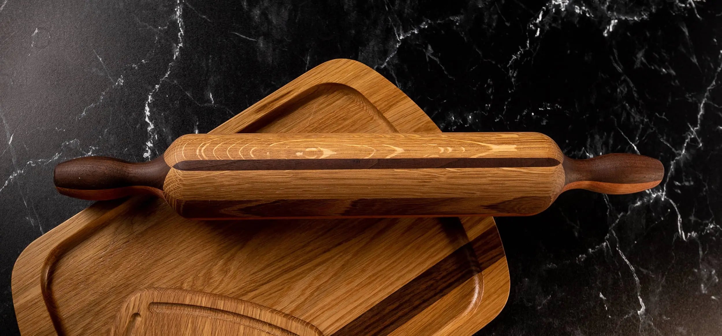 End grain rolling pin with dark and light wood patterns, resting on a wooden cutting board, displayed on a black marble surface. High-quality kitchen tool for baking and cooking.