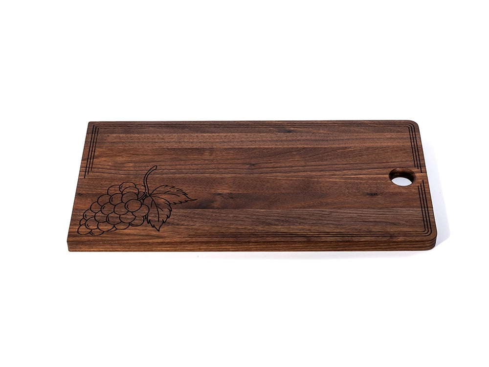 Black walnut charcuterie board with laser-engraved grape design, decorative border, and hanging hole.