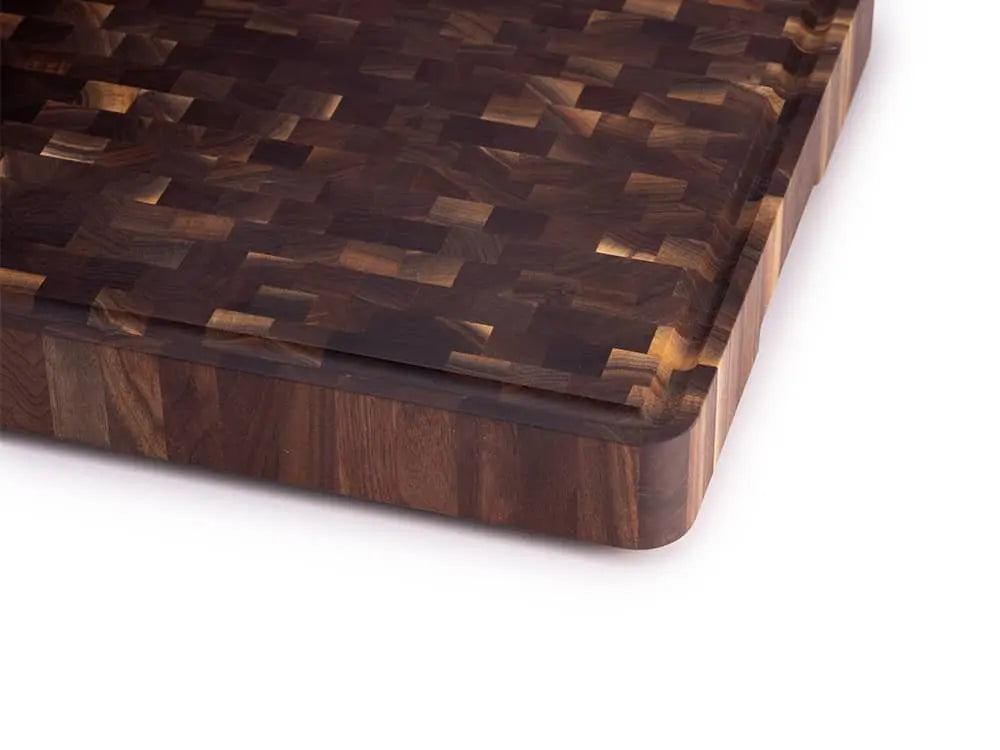 Close-up of a black walnut end grain butcher block corner with a checkered pattern, handle cutout, and black rubber foot, against a white background