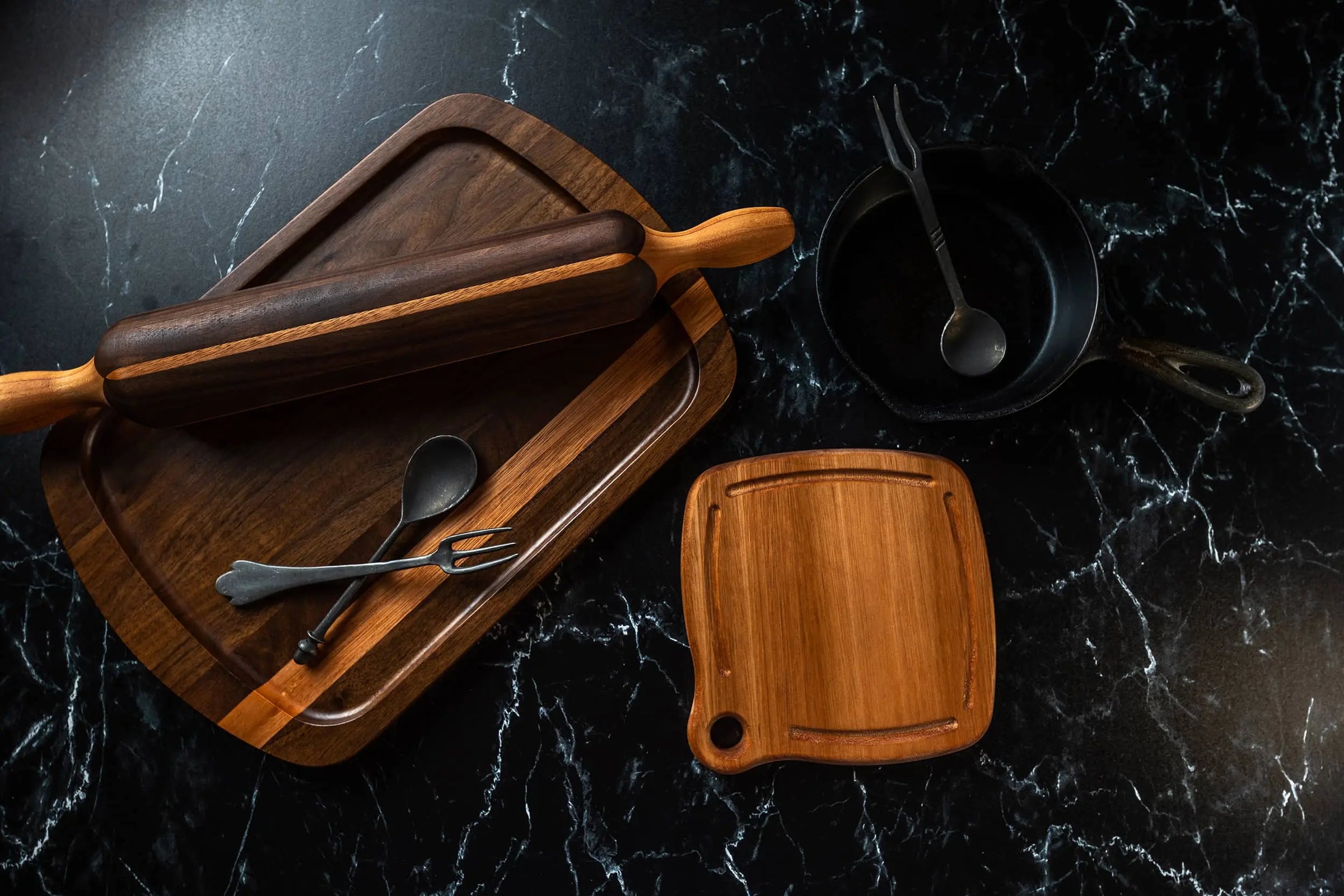 Kitchen essentials displayed on a dark marble countertop, featuring a wooden rolling pin, various wooden cutting boards, metal utensils, and a cast iron skillet