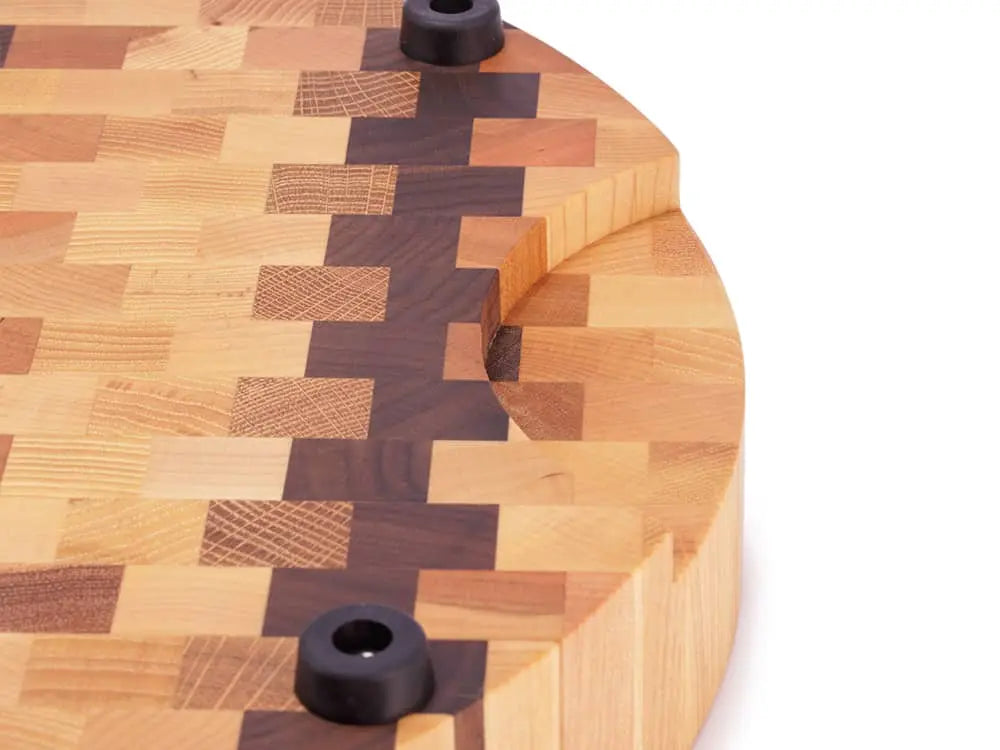 Close-up of a round end grain butcher block with a checkered pattern, showing a handle cutout and black rubber feet, against a white background